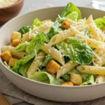 A fresh Caesar pasta salad in a beige bowl, featuring penne pasta, crisp romaine lettuce, golden croutons, and shredded Parmesan cheese, set on a light gray table with a fork and grater nearby.